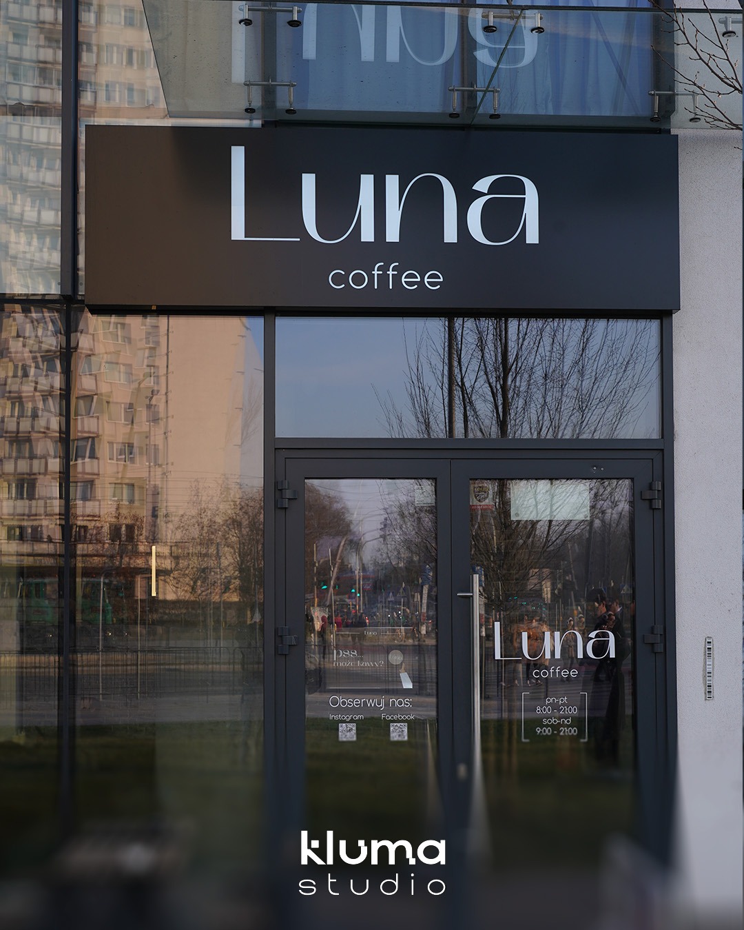 Zewnętrzny szyld z logo 'Luna coffee' wykonany z błyszczącego czarnego Dibondu, podświetlony zimnym światłem LED, zamontowany nad wejściem do kawiarni z przeszklonymi drzwiami, odbijającymi ruch...