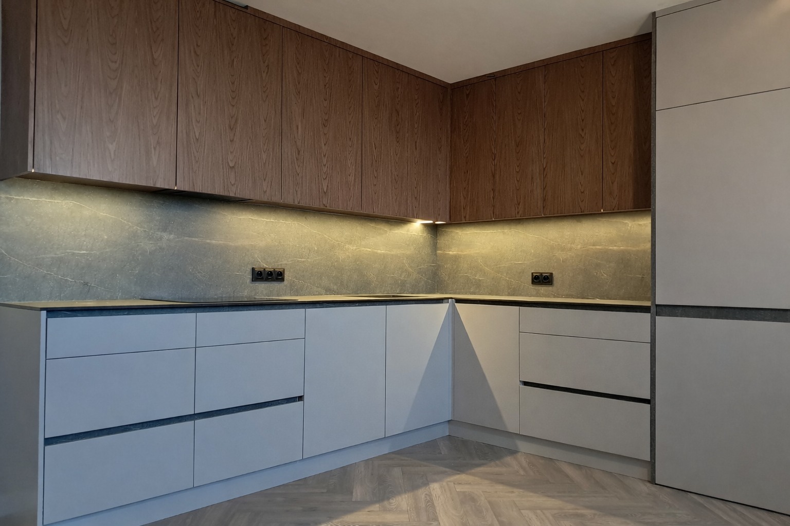 Nowoczesna kuchnia w odcieniach szarości i drewna z subtelnym oświetleniem LED pod szafkami. Widoczny parkiet ułożony w jodełkę i gładkie fronty szafek.