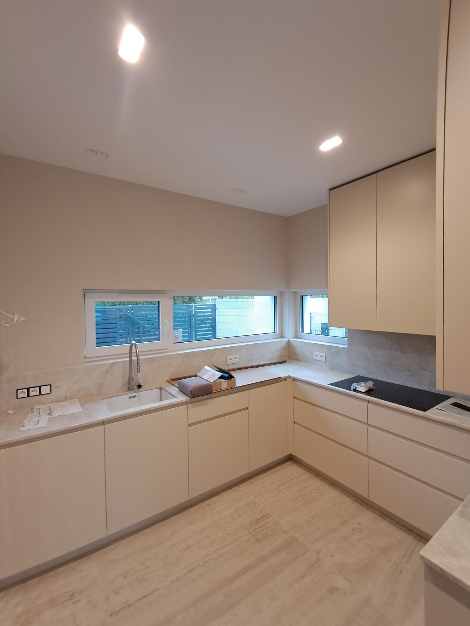 Jasna, nowoczesna kuchnia z beżowymi szafkami, marmurowym blatem i podłogą. Widoczne okno z widokiem na zewnątrz i minimalistyczna armatura.