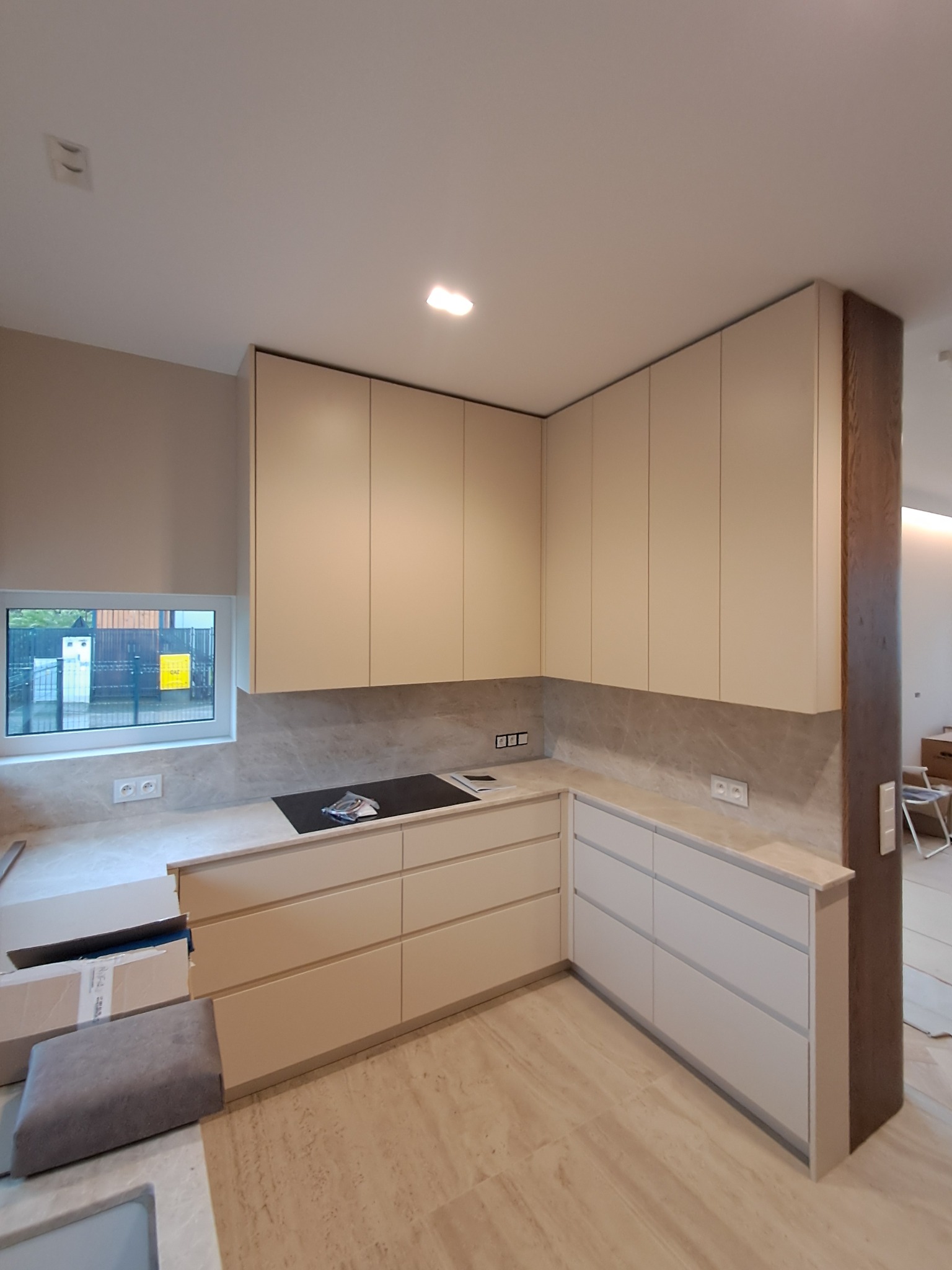 Jasna, nowoczesna zabudowa kuchenna z beżowymi frontami i marmurowym blatem. Widoczne gniazdka elektryczne i płyta indukcyjna. Częściowo widoczne kartony po montażu.