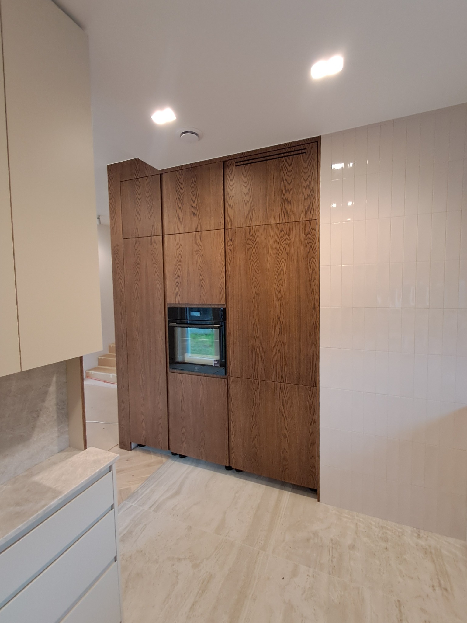 Nowoczesna zabudowa kuchenna z drewnianymi frontami i wbudowanym piekarnikiem, jasne płytki na ścianie, marmurowa podłoga. Widok na schody w tle.