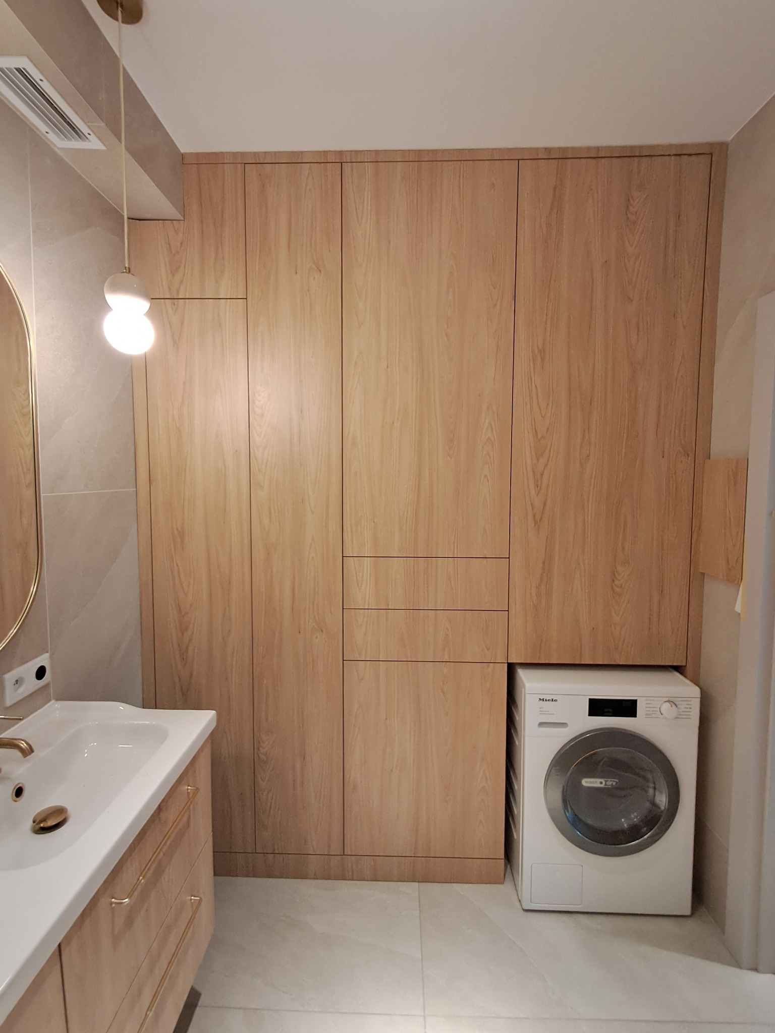 Jasne, drewniane szafy w zabudowie w łazience, zintegrowane z umywalką i pralką Miele. Minimalistyczny design, ciepłe oświetlenie.