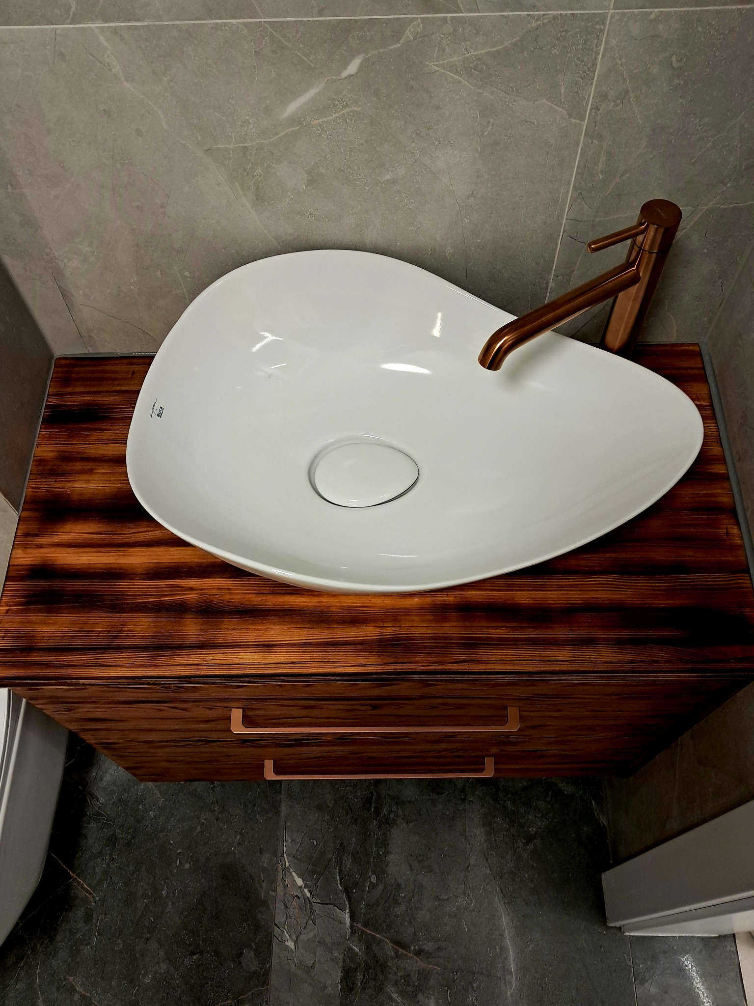 Umywalka ceramiczna o nieregularnym kształcie, osadzona na szafce z ciemnego drewna z widocznymi usłojeniami, bateria w kolorze miedzi, szare płytki w tle.