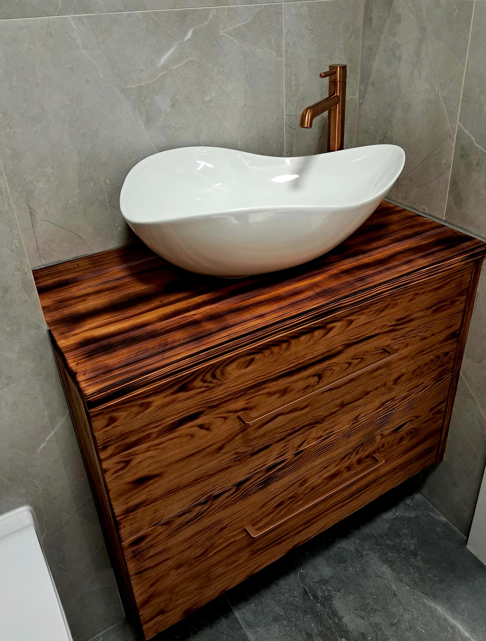 Nowoczesna szafka łazienkowa z palonego drewna, umywalką w kształcie łzy i miedzianą armaturą na tle szarej marmurowej ściany.