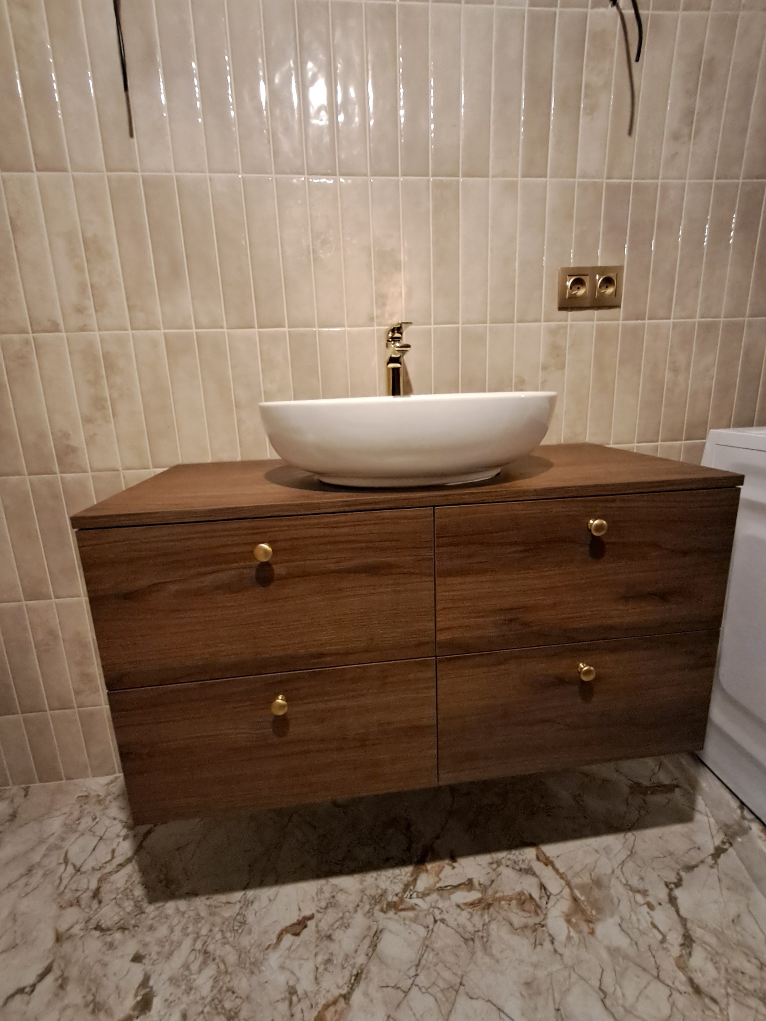 Nowoczesna szafka łazienkowa z umywalką nablatową i złotą armaturą na tle beżowych płytek. Widoczne marmurowe płytki podłogowe i gniazdka elektryczne.