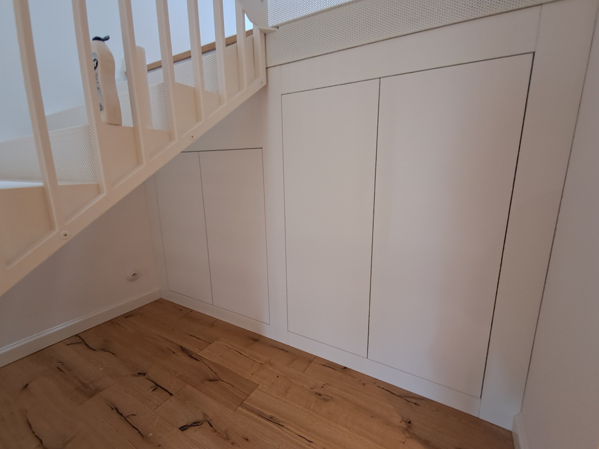 Białe szafki wnękowe pod schodami, drewniana podłoga. Minimalistyczny design, praktyczne wykorzystanie przestrzeni. Widok z dołu, jasne oświetlenie.