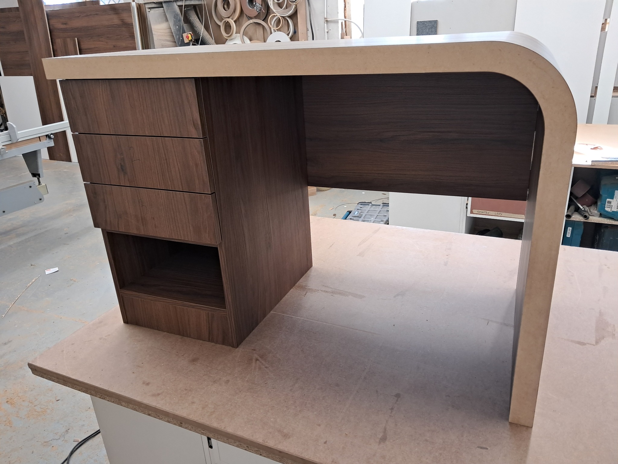 Nowoczesne biurko z ciemnego drewna z trzema szufladami i półką, z zaokrąglonym blatem, w warsztacie stolarskim. Widoczne narzędzia i materiały w tle.