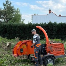 Oleksandr Kotov, Warszawski Arborysta - Pracownik w kombinezonie obsługuje pomarańczową rębarkę Timberwolf TW 230DHB na tle zieleni i białego domu z kominem. Widoczne ścięte pnie drzew.