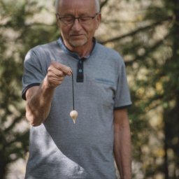 Gabinet Naturoterapii - Mężczyzna z wahadełkiem na tle zieleni, skupiony wzrok, sesja z wahadłem w otoczeniu natury, jasne drewno wahadełka.