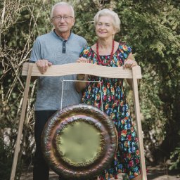 Gabinet Naturoterapii - Para seniorów pozuje z gongiem na drewnianym stelażu w plenerze. Kobieta w kwiecistej sukience, mężczyzna w szarej koszulce. Tło z krzewów i drzew.