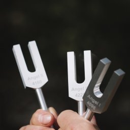 Gabinet Naturoterapii - Dłoń trzymająca trzy kamertony 'Angel' o różnych częstotliwościach na ciemnym tle. Narzędzia do terapii dźwiękiem w medycynie naturalnej.