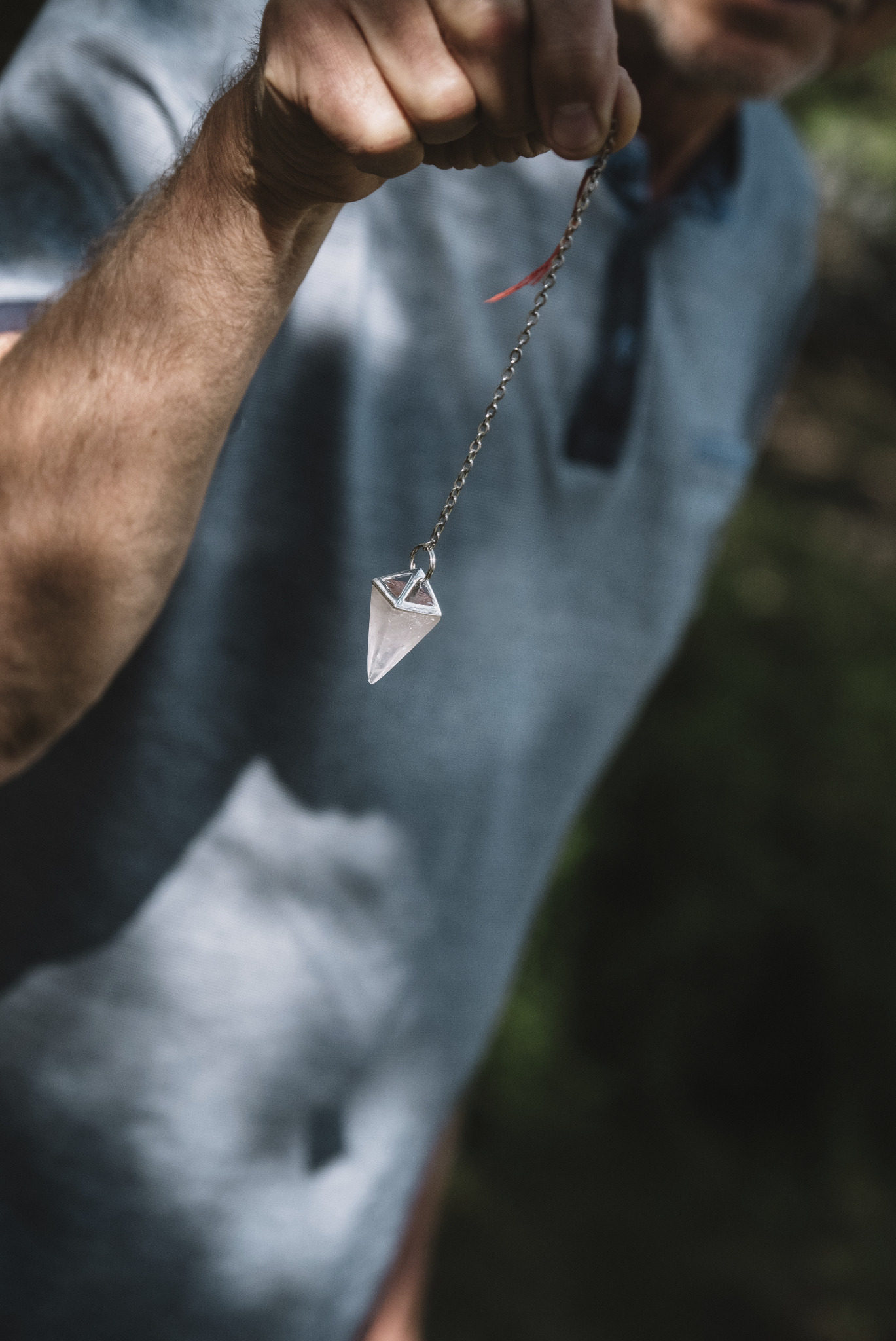 Kryształowy wahadełko na łańcuszku, trzymane w dłoni, w tle zamazane ubranie i zieleń. Narzędzie używane w radiestezji i medycynie holistycznej.