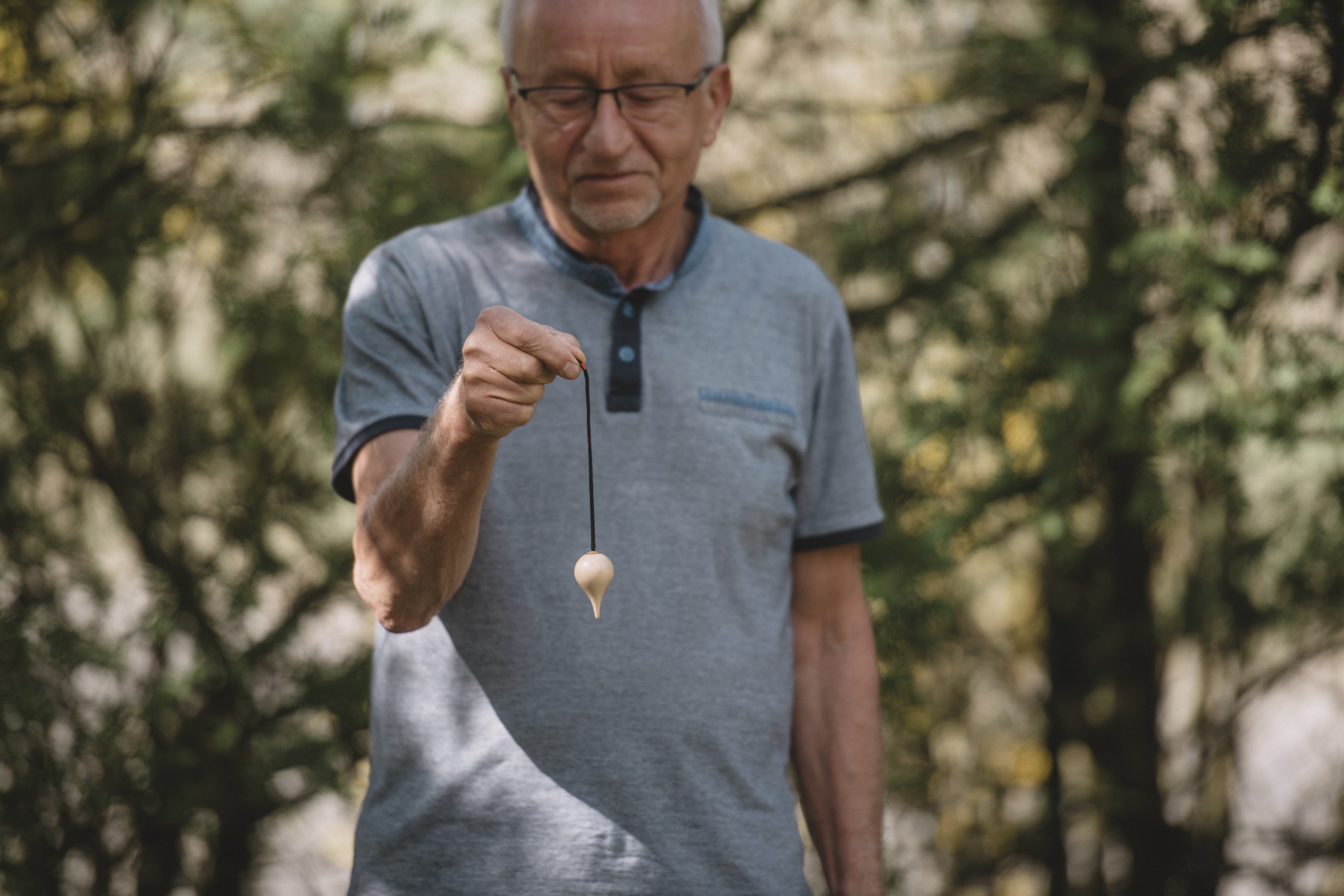 Mężczyzna z wahadełkiem na tle zieleni, skupiony wzrok, sesja z wahadłem w otoczeniu natury, jasne drewno wahadełka.