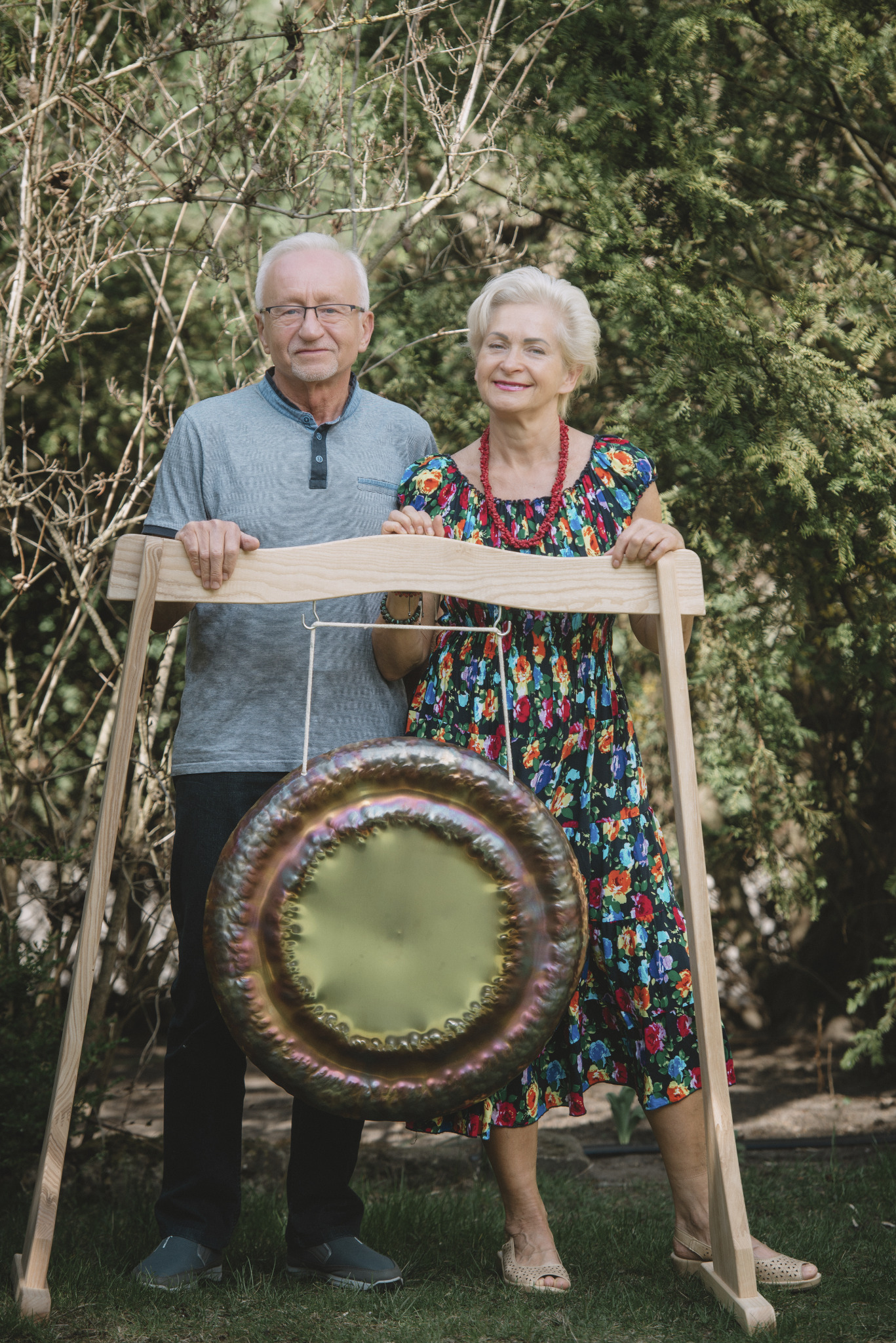 Para seniorów pozuje z gongiem na drewnianym stelażu w plenerze. Kobieta w kwiecistej sukience, mężczyzna w szarej koszulce. Tło z krzewów i drzew.