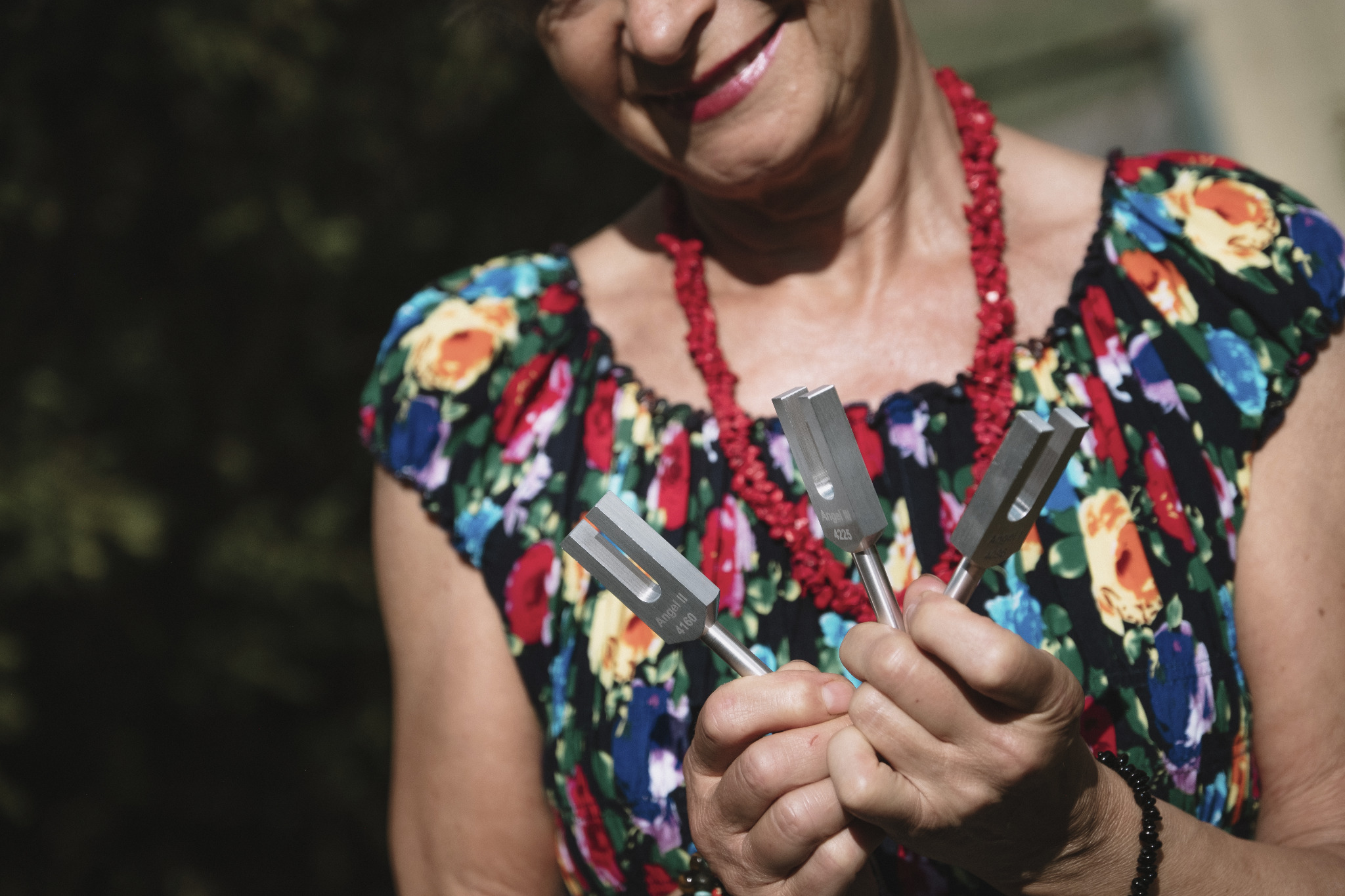 Kobieta z uśmiechem trzymająca w dłoniach trzy kamertony, narzędzia używane w terapii dźwiękiem. Widoczny kwiecisty strój i czerwone korale.