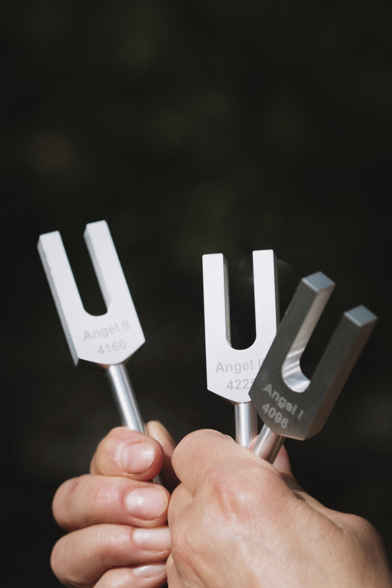 Dłoń trzymająca trzy kamertony 'Angel' o różnych częstotliwościach na ciemnym tle. Narzędzia do terapii dźwiękiem w medycynie naturalnej.