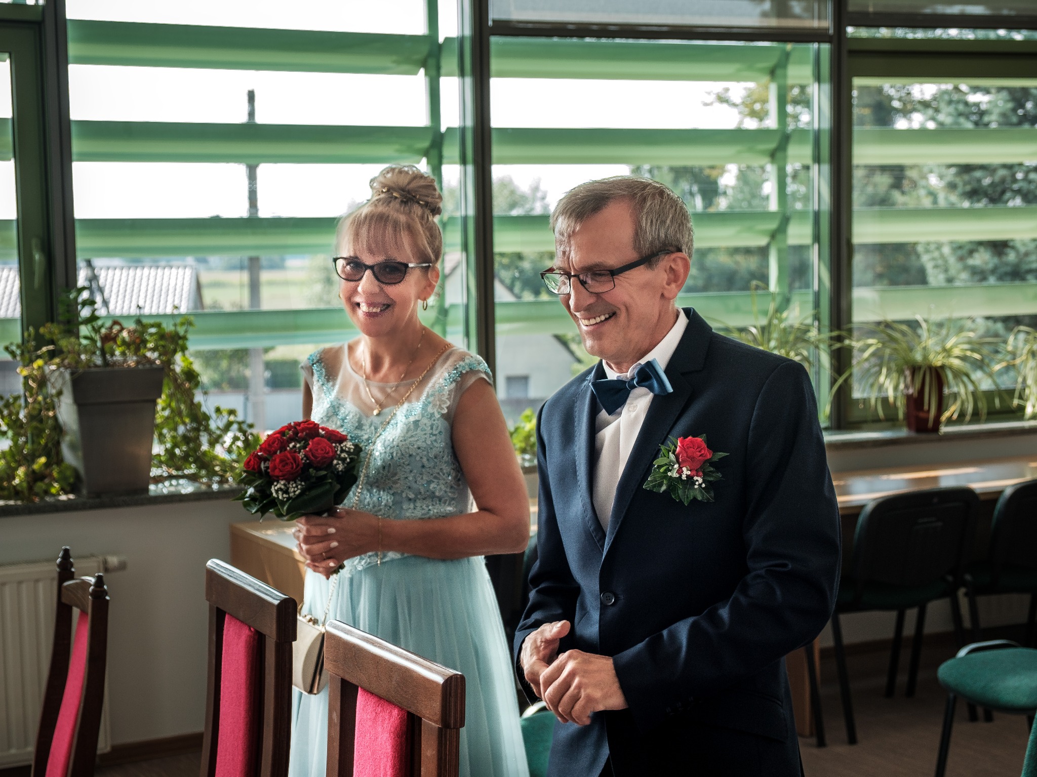 Uśmiechnięta para w strojach ślubnych, panna młoda z bukietem czerwonych róż, elegancki pan młody w ciemnym garniturze z muszką, wnętrze z dużymi oknami.