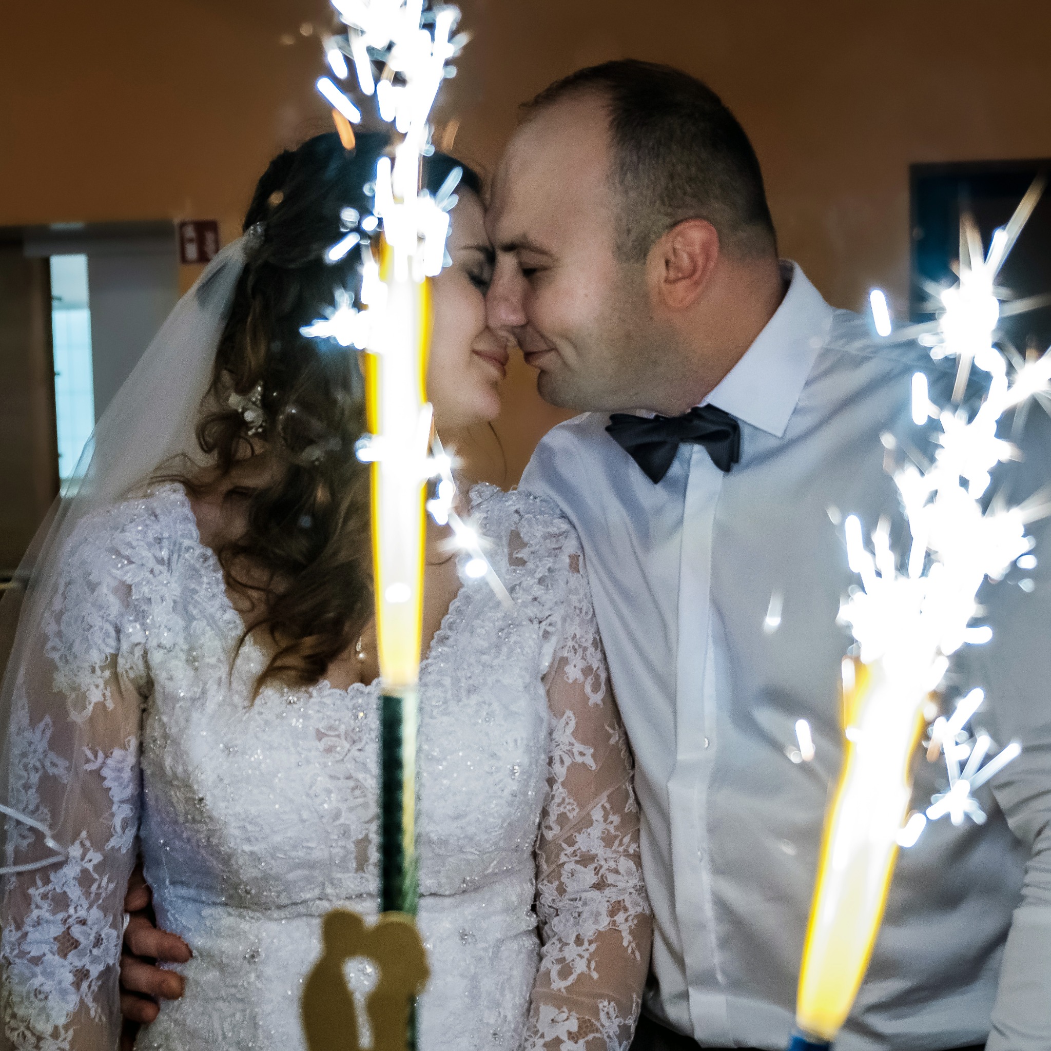 Para młoda w zbliżeniu, z iskrzącymi fontannami tortowymi. Panna młoda w koronkowej sukni, pan młody w koszuli i muszce. Romantyczna atmosfera.