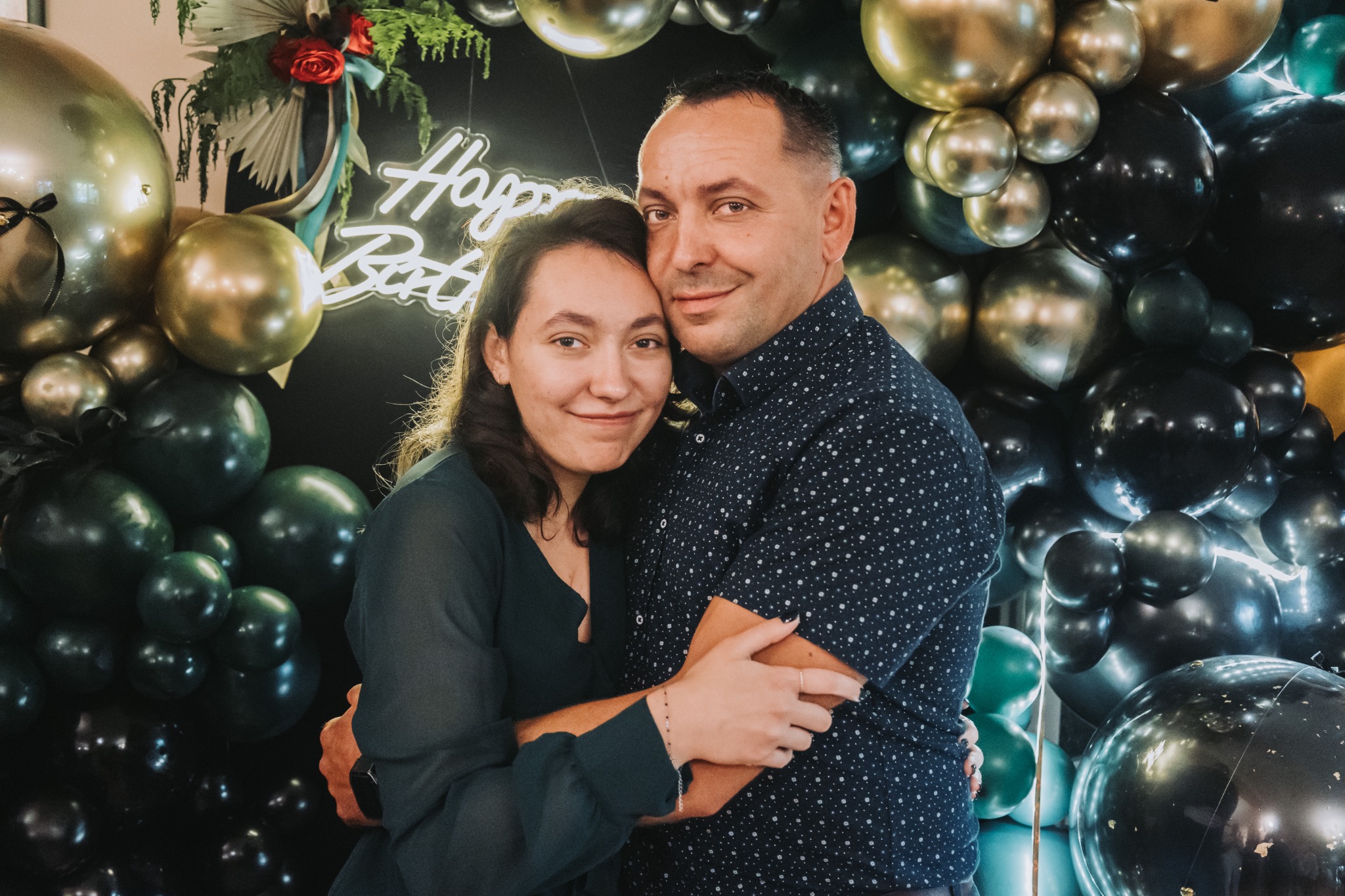 Uśmiechnięta para obejmuje się na tle dekoracji z balonów i neonu 'Happy Birthday'. Zdjęcie z imprezy urodzinowej, wykonane w ciemnej tonacji.