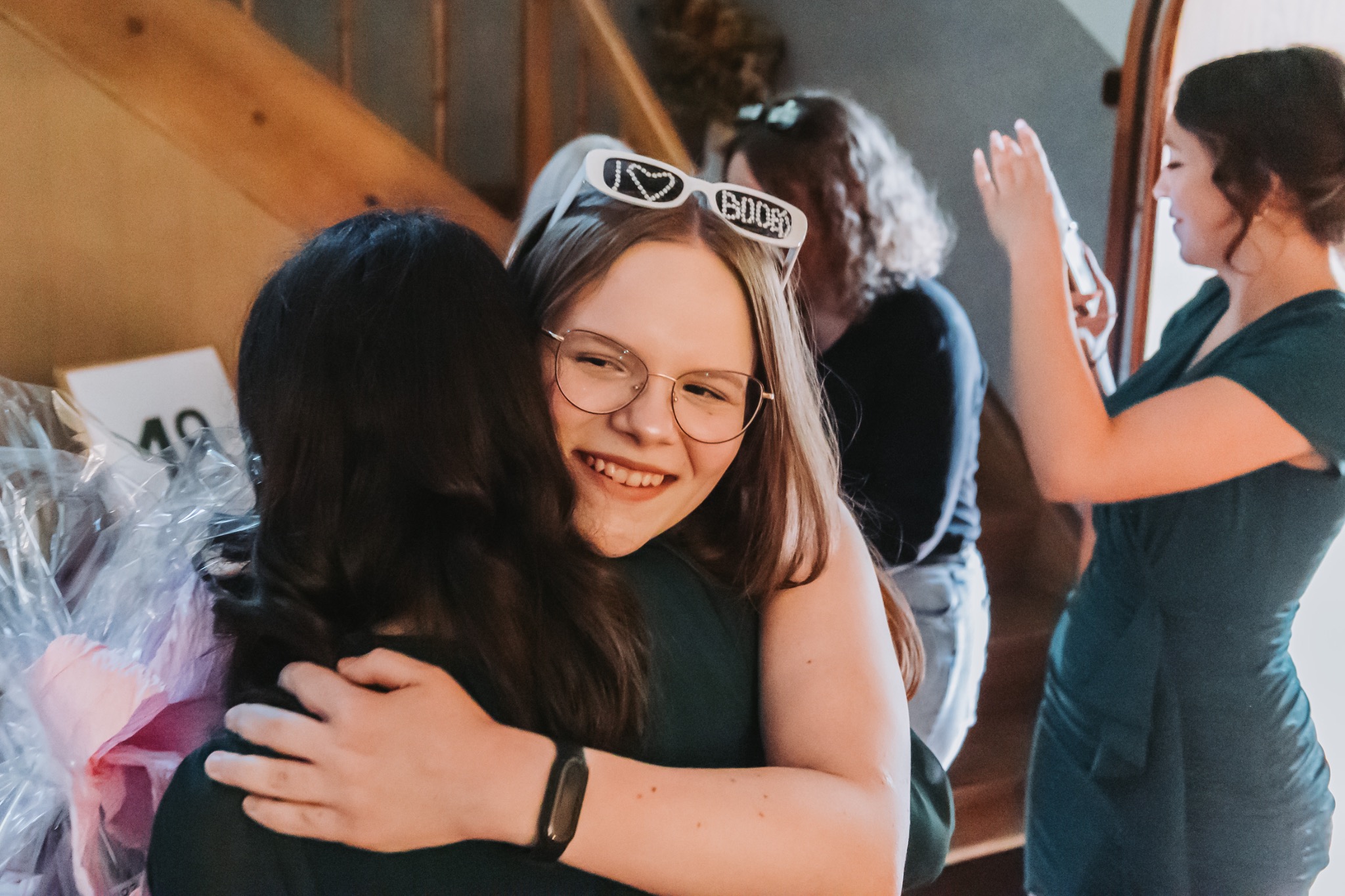 Uśmiechnięta dziewczyna w okularach z napisem 'I love boobies' obejmuje kogoś podczas imprezy. W tle inne osoby i schody.