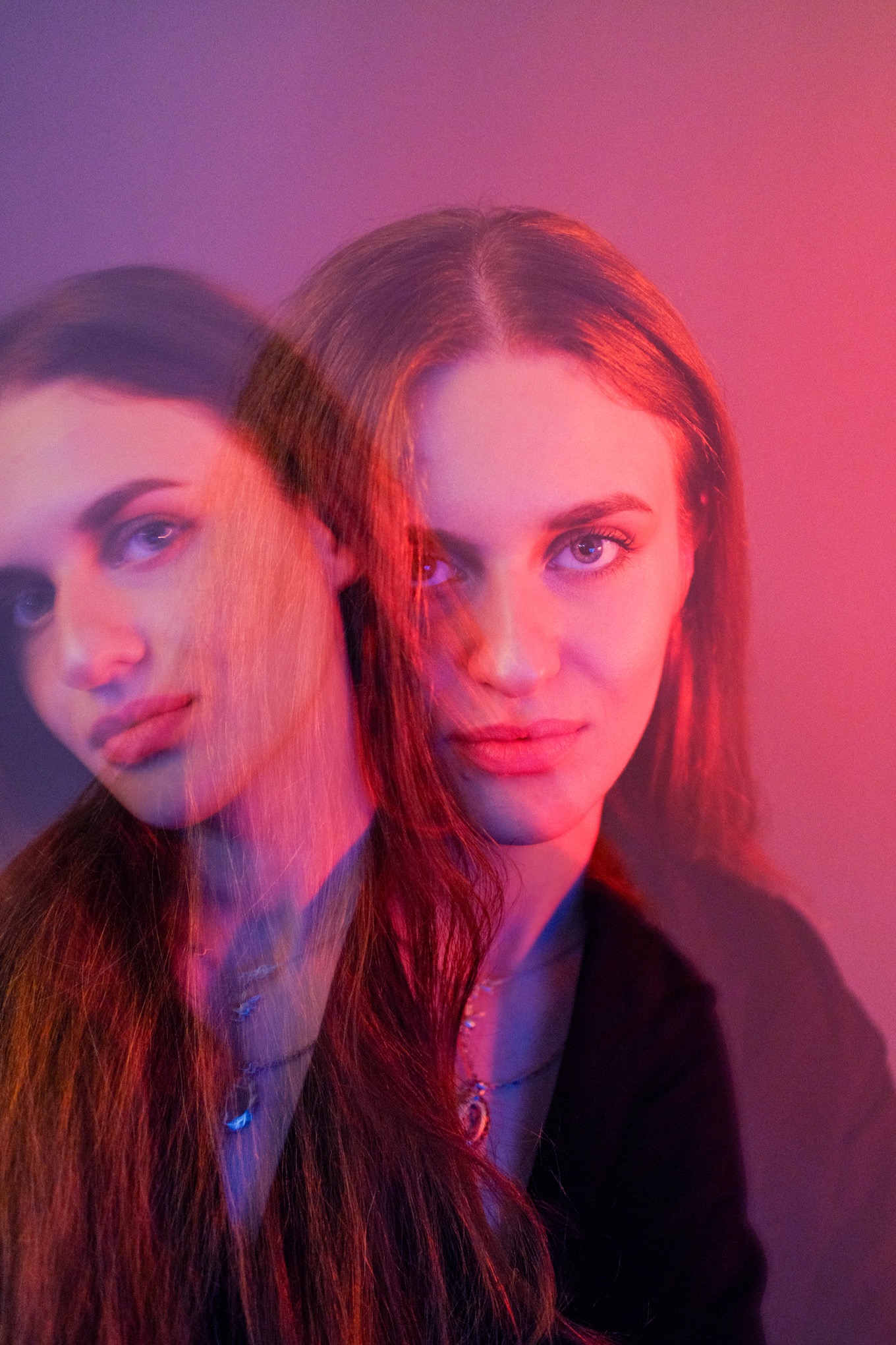 Portret kobiety z efektem podwójnej ekspozycji w purpurowo-różowym oświetleniu. Widoczne srebrne naszyjniki i ciemne ubranie. Artystyczne ujęcie.