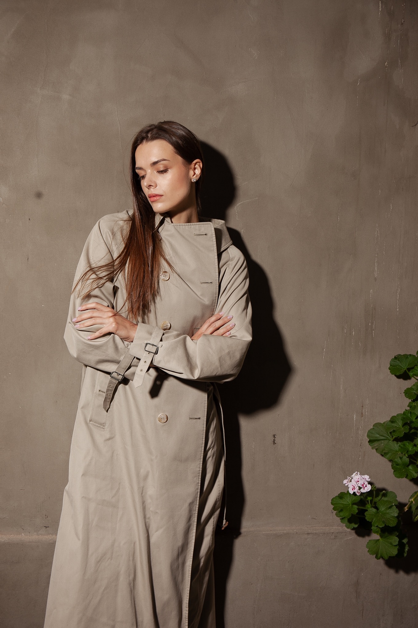 Portret kobiety w beżowym płaszczu na tle szarej ściany i zielonej rośliny. Ujęcie z delikatnym światłem, podkreślające naturalność i elegancję.