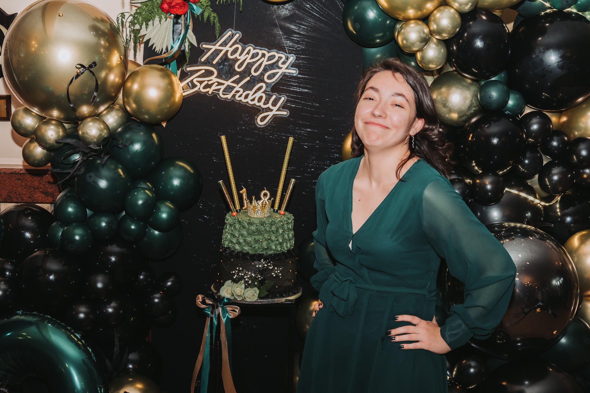 Uśmiechnięta kobieta w szmaragdowej sukience pozuje obok tortu urodzinowego z dekoracją balonową w odcieniach złota i czerni. Neonowy napis 'Happy Birthday'.