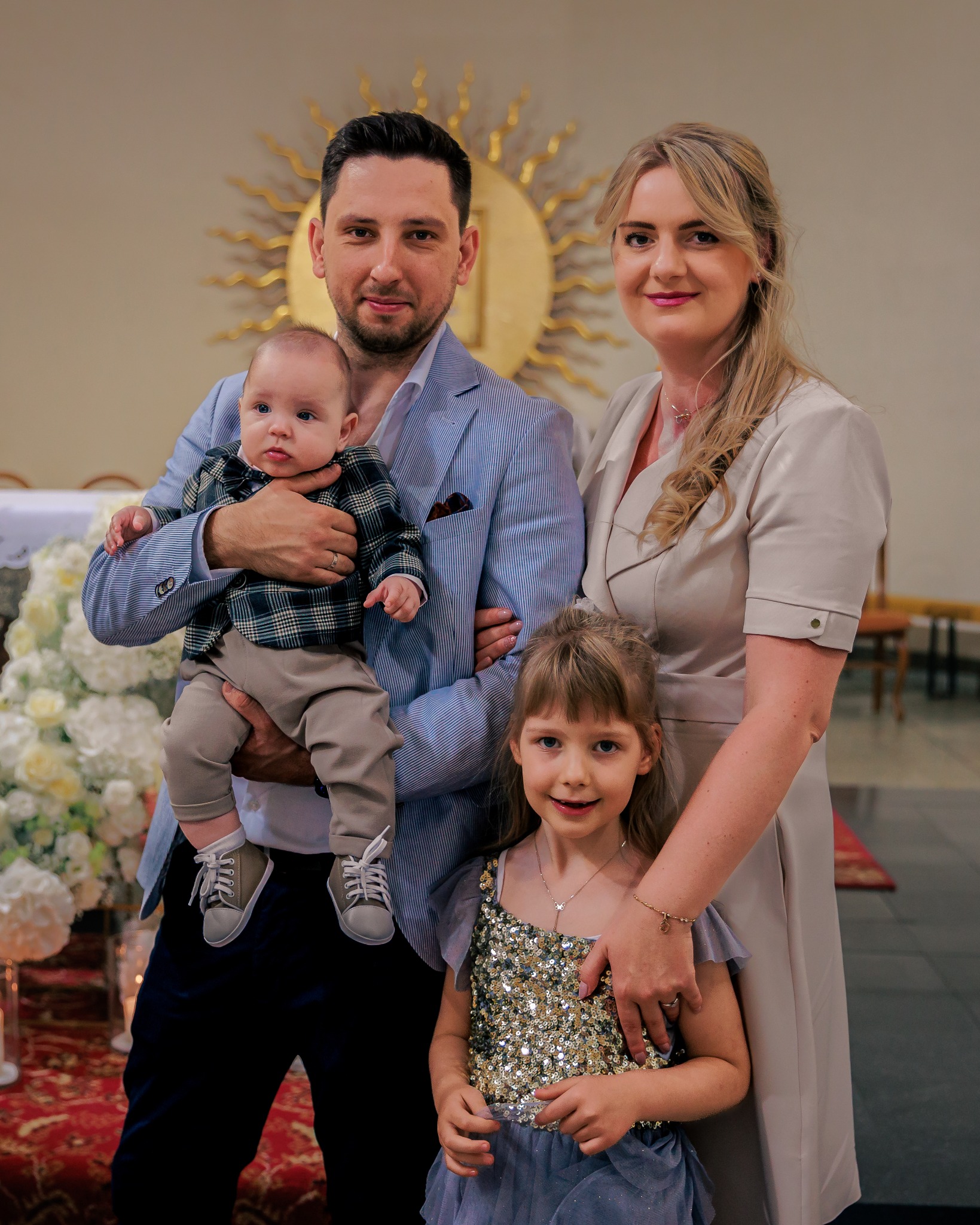 Uśmiechnięta rodzina w kościele: tata trzyma synka w eleganckiej stylizacji, mama i córka obok. W tle ołtarz z kwiatami i złote słońce.
