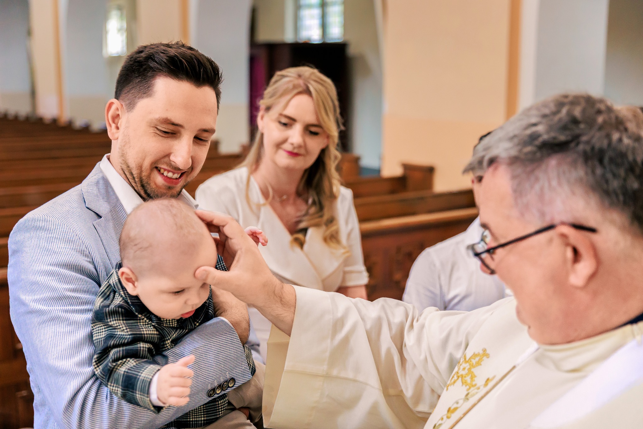 Uśmiechnięty ojciec trzyma dziecko w trakcie chrztu, na tle matka i ksiądz w kościele. Jasne wnętrze, delikatne światło, uroczysta atmosfera.