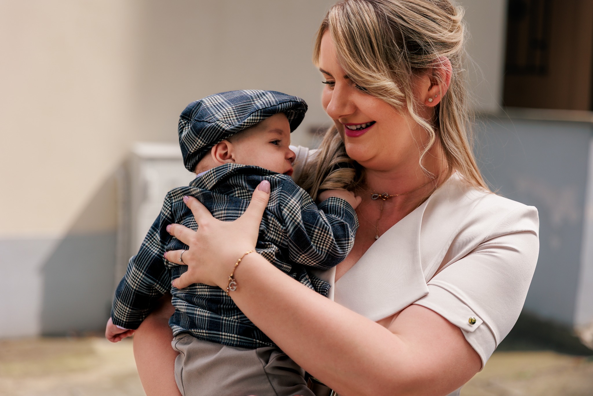 Uśmiechnięta mama trzymająca na rękach elegancko ubranego synka w czapce w kratę. Delikatny makijaż i biżuteria kobiety. Jasne tło.