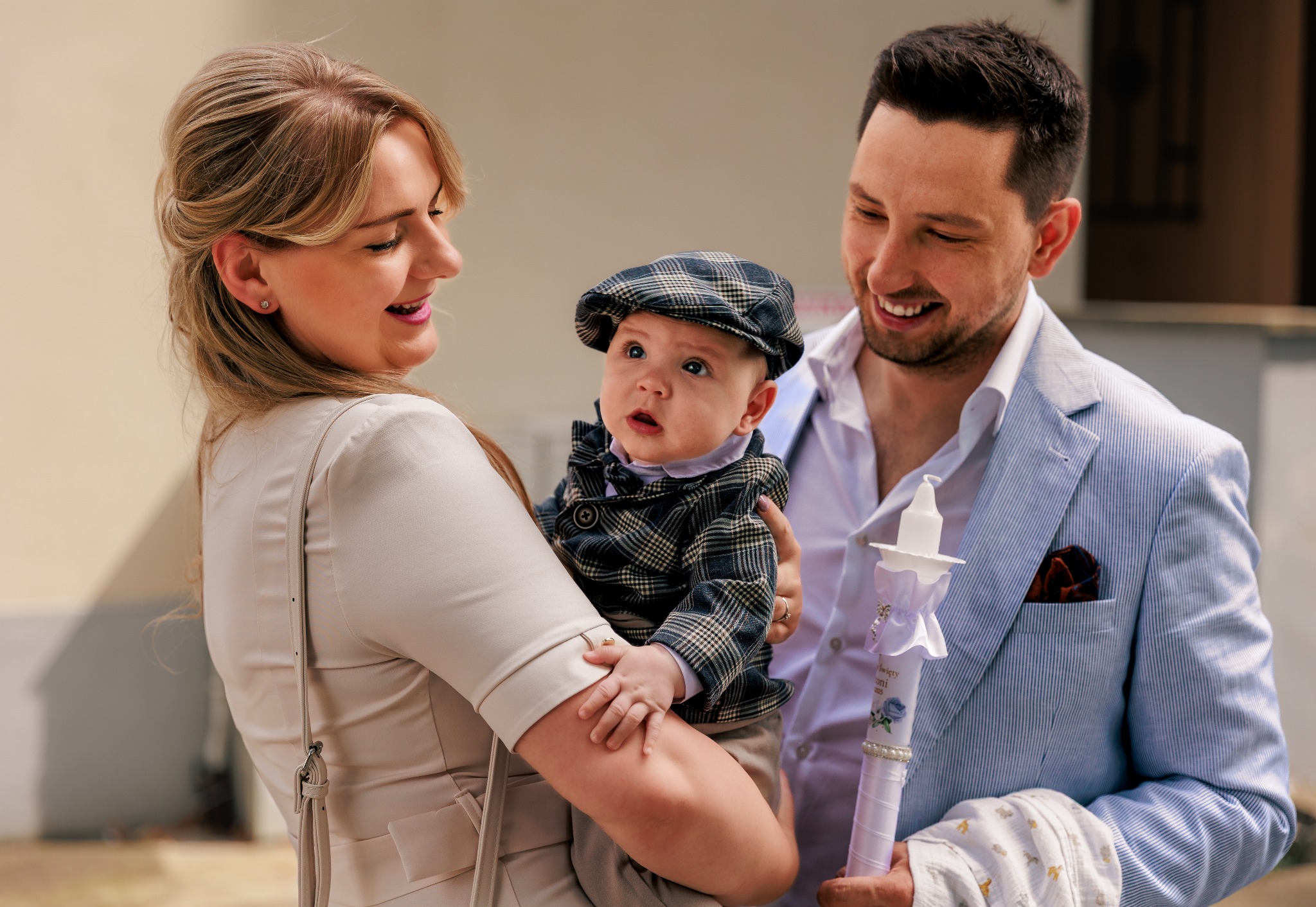 Uśmiechnięta rodzina z dzieckiem w czapce kaszkietówką i świecą do chrztu. Mama trzyma dziecko, tata obok. Jasne, naturalne światło.