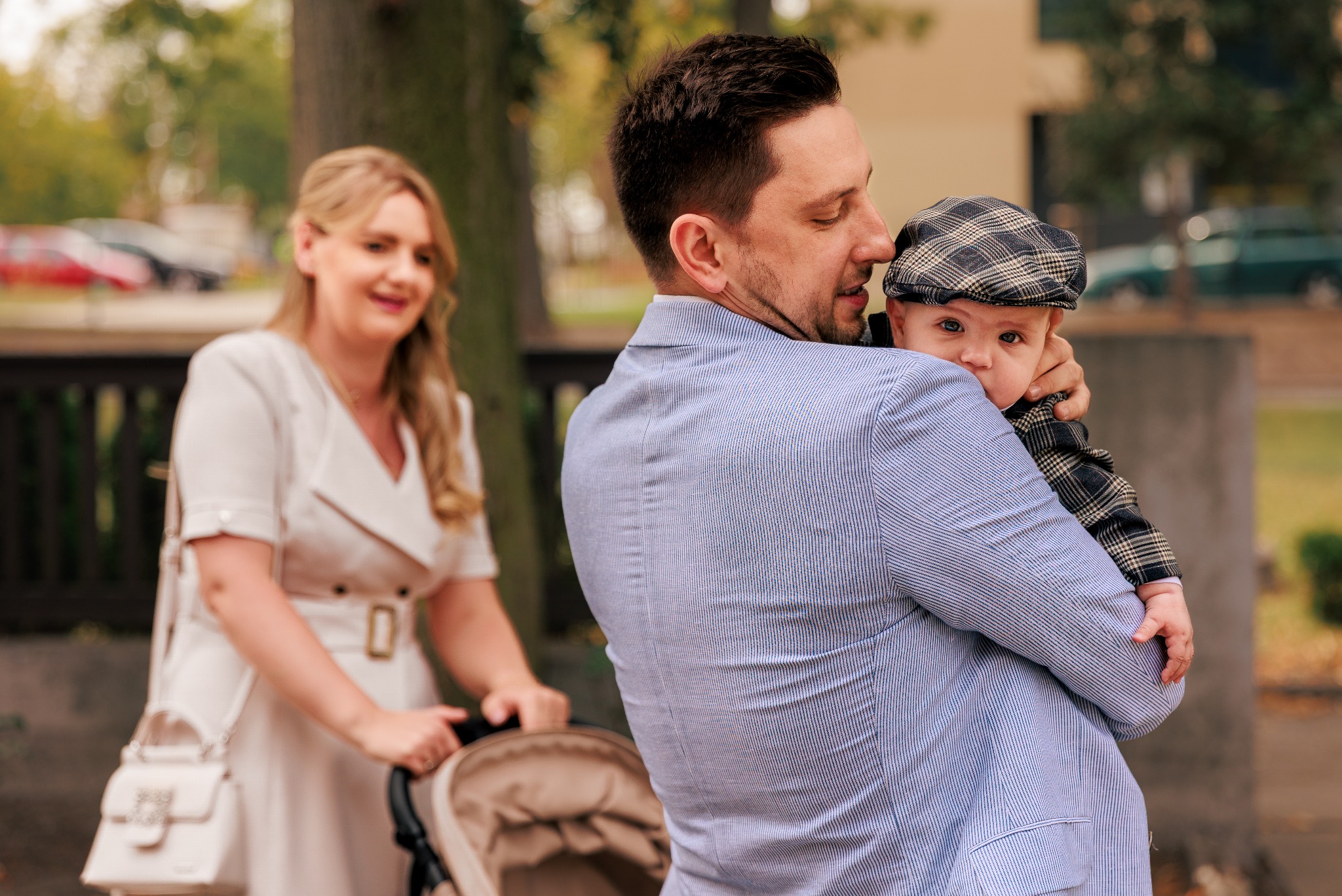 Uśmiechnięty ojciec trzymający na rękach synka w czapce, w tle mama z wózkiem. Sesja rodzinna w parku, jesienna sceneria, naturalne światło.