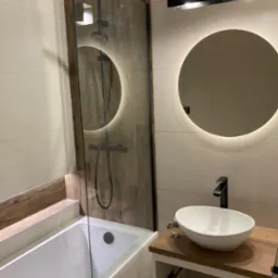 Nowoczesna łazienka z wanną za szklaną ścianką, deszczownicą w kolorze czarnym, okrągłym lustrem z podświetleniem LED i umywalką nablatową na drewnianym blacie.