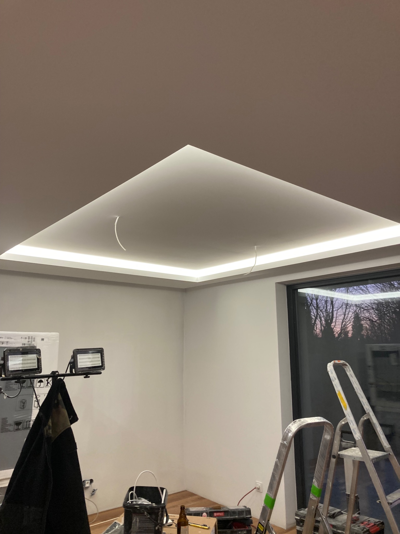 Nowoczesne oświetlenie sufitowe z listwami LED w trakcie montażu, widoczne kable i drabiny, narzędzia pracy na podłodze.