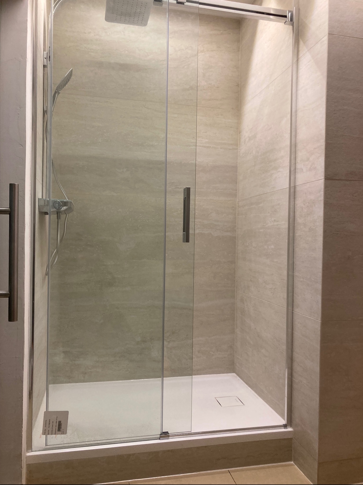Kabina prysznicowa z deszczownicą i słuchawką, wykończona płytkami imitującymi trawertyn, z widoczną etykietą informacyjną na szybie.