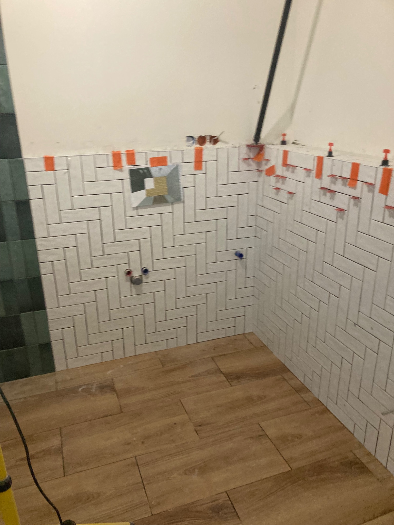 Łazienka w trakcie remontu: białe, ułożone w jodełkę płytki na ścianach z pomarańczowymi elementami poziomowania, drewnopodobne płytki na podłodze, widoczne rury i otwory na armaturę, fragment...