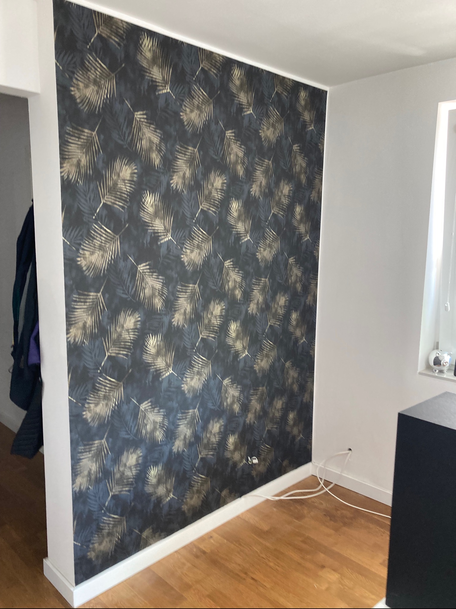 Ściana w pokoju po remoncie z tapetą w ciemne liście z elementami złota, widoczne gniazdko elektryczne i listwa przypodłogowa.