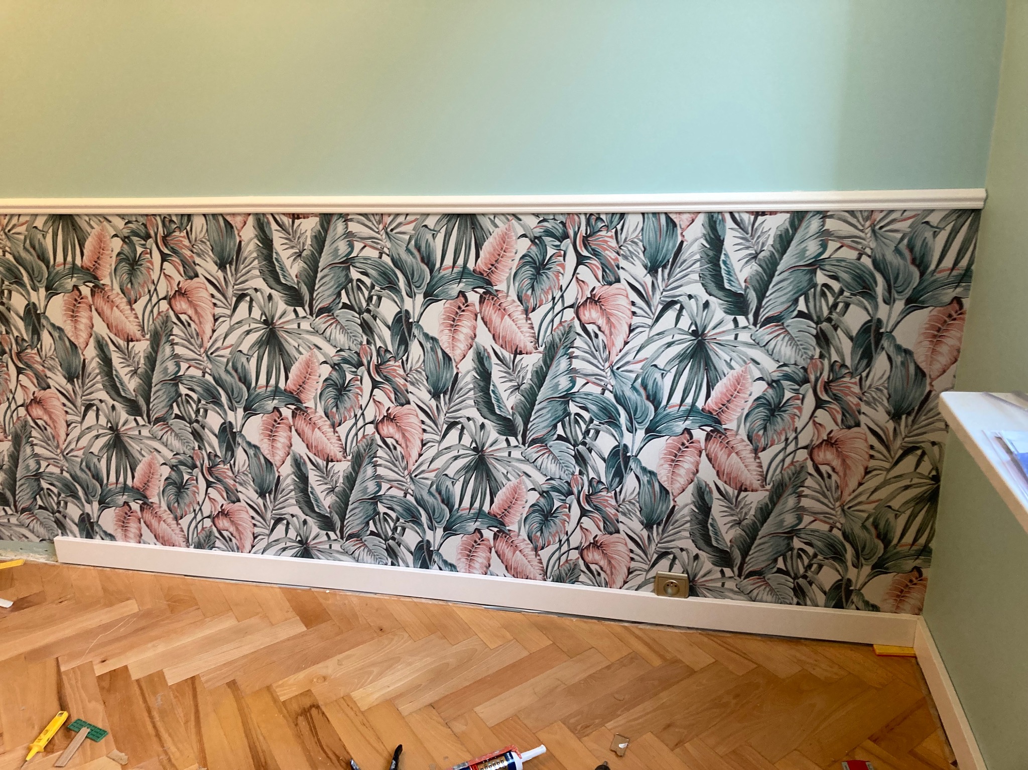 Ściana po remoncie z nową tapetą w tropikalne liście, listwa przypodłogowa zamontowana, widoczne narzędzia na parkiecie w jodełkę.
