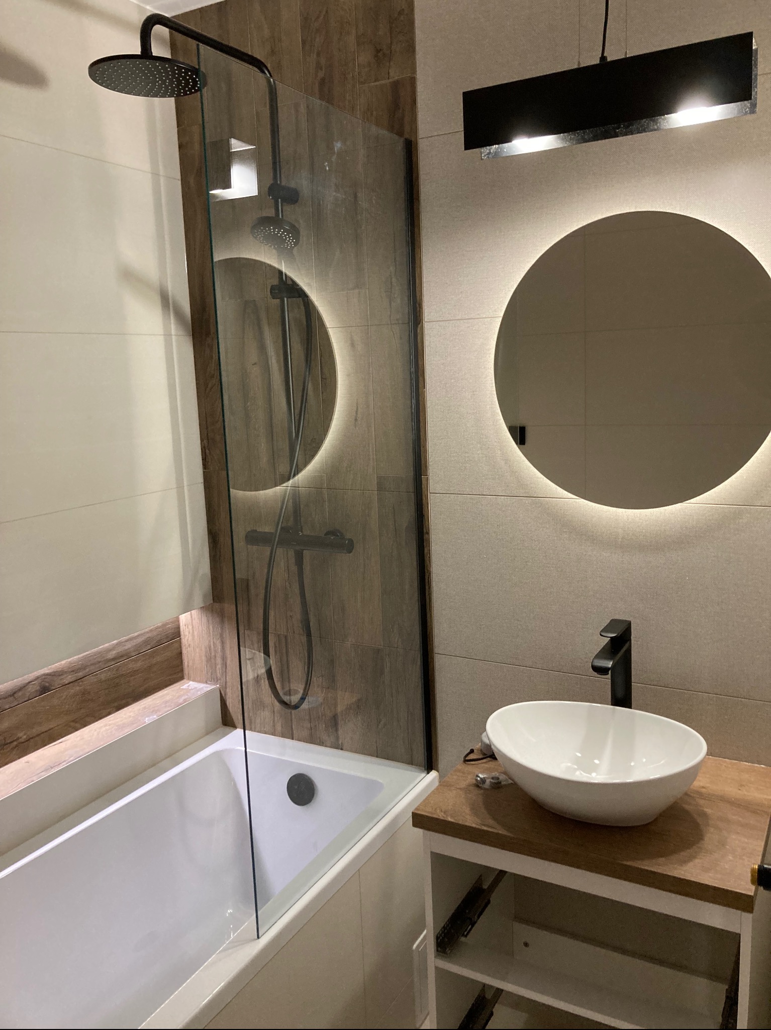 Nowoczesna łazienka z wanną za szklaną ścianką, deszczownicą w kolorze czarnym, okrągłym lustrem z podświetleniem LED i umywalką nablatową na drewnianym blacie.