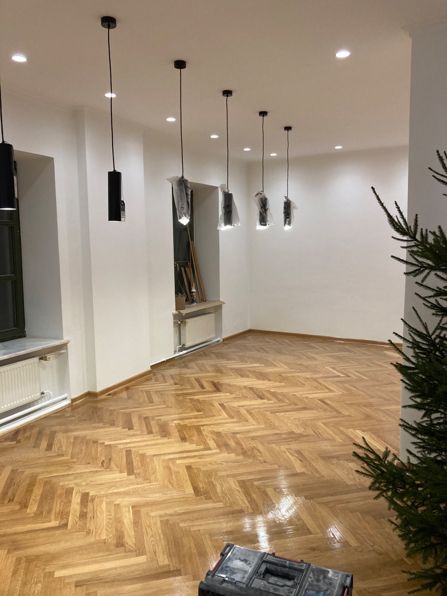 Pusty pokój po remoncie z parkietem w jodełkę, świeżo pomalowanymi białymi ścianami, nowymi czarnymi lampami wiszącymi jeszcze w folii ochronnej i narzędziami w skrzynce na podłodze.