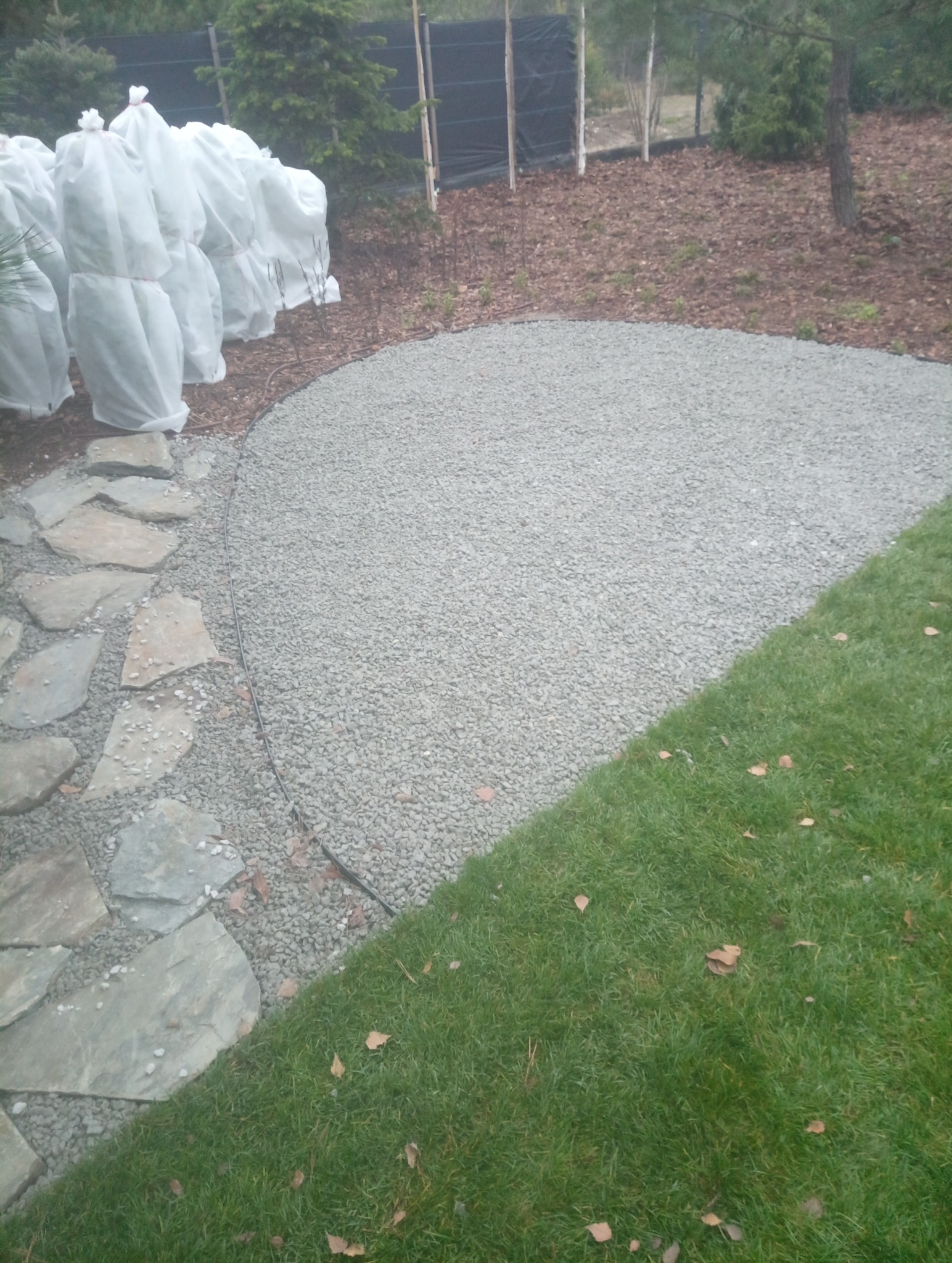 Kaskada kamiennych płyt przechodzi w szarą, żwirową ścieżkę, obok której rośnie trawa. W tle widoczne drzewa, niektóre zabezpieczone białym materiałem.