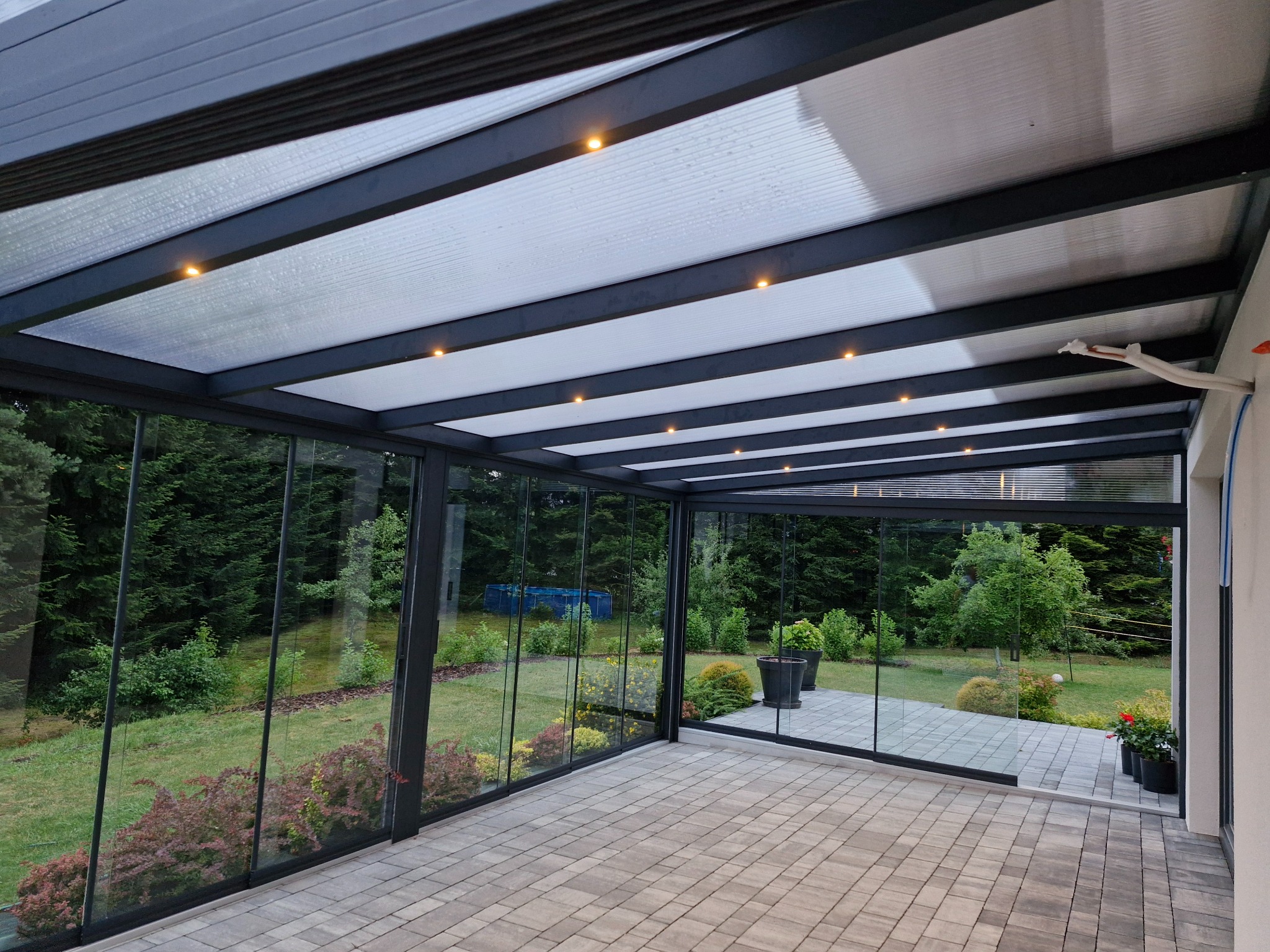 Zadaszony taras z metalową konstrukcją i szklanymi ścianami. Widok na ogród, kostka brukowa na podłodze, oświetlenie punktowe w suficie.