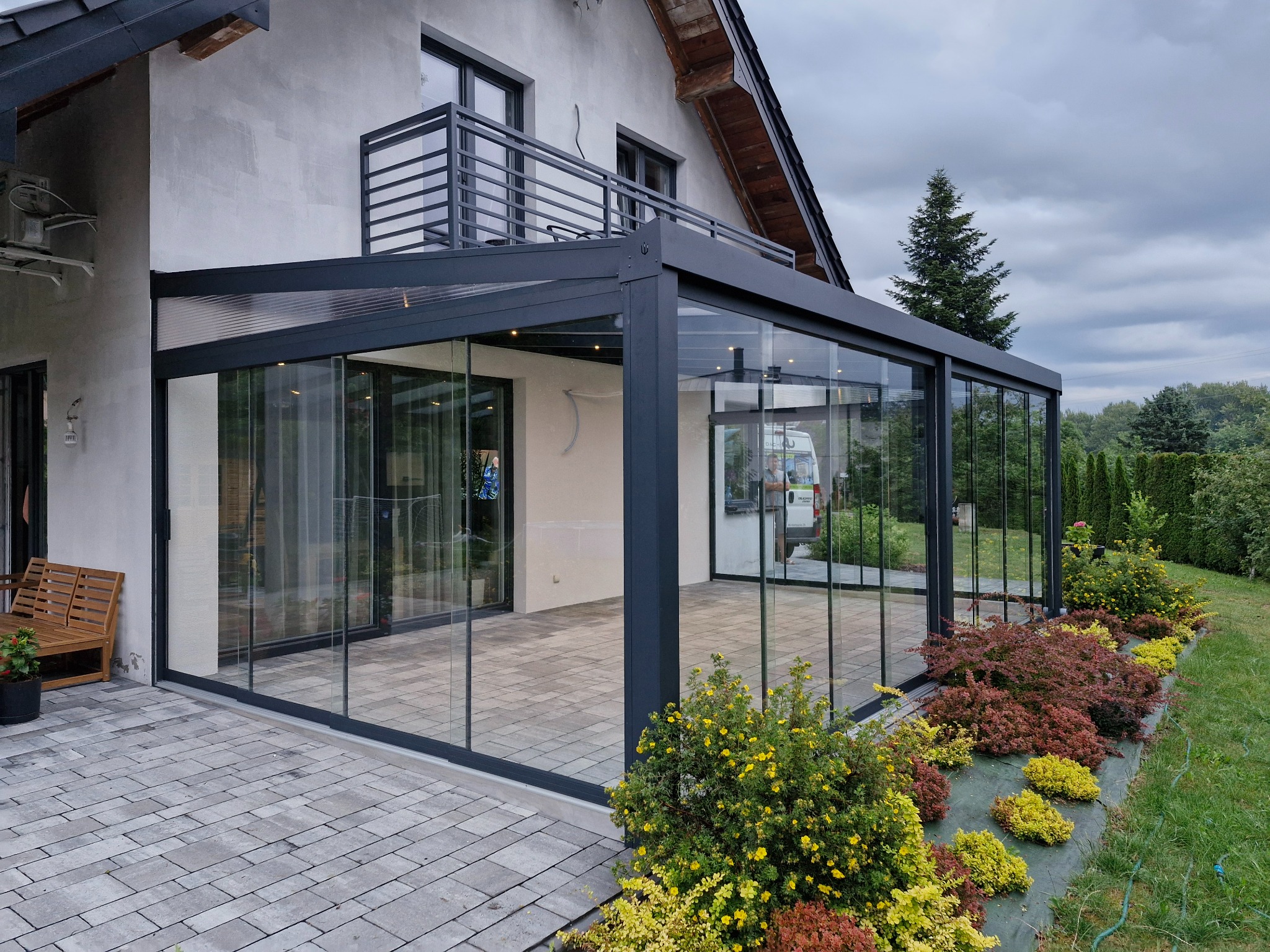 Szklana zabudowa tarasu z metalową konstrukcją, przylegająca do domu z balkonem. Widoczna kostka brukowa i ozdobne krzewy. Całość w neutralnej kolorystyce.