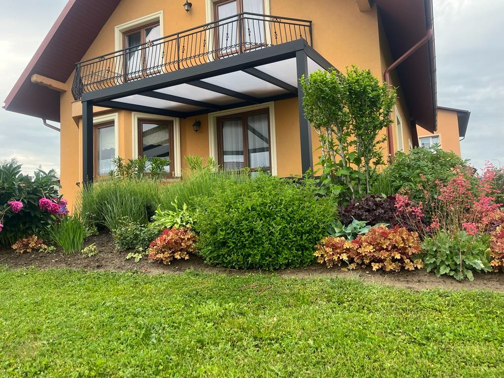 Zadaszenie tarasu z czarną, metalową konstrukcją i transparentnym dachem, przylegające do domu z balkonem. Uwagę przyciąga bujna, kolorowa roślinność w ogrodzie.