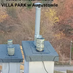 Szczegółowe ujęcie kominów z obróbkami blacharskimi na dachu budynku z widokiem na jesienne drzewa w tle. Widoczne elementy uszczelniające i rury wentylacyjne.