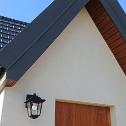 USŁUGI OGÓLNOBODOWLANE -ALEXANDRU ANTON - Fragment domu z ciemną dachówką, rynnami i obróbkami blacharskimi. Widoczne oświetlenie zewnętrzne i fragment drzewa na tle błękitnego nieba.