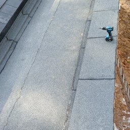 USŁUGI OGÓLNOBODOWLANE -ALEXANDRU ANTON - Widok z góry na fragment dachu z papy termozgrzewalnej, na którym leży wkrętarka akumulatorowa. W tle dachówka i drabina oparta o ścianę budynku.