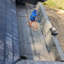 USŁUGI OGÓLNOBODOWLANE -ALEXANDRU ANTON - Dekarz pracuje na dachu z czarnej dachówki, używając palnika podłączonego do niebieskiej butli gazowej. Widoczne narzędzia i elementy konstrukcji dachu.