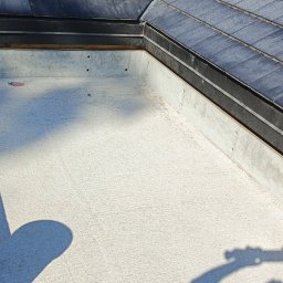 USŁUGI OGÓLNOBODOWLANE -ALEXANDRU ANTON - Betonowa attyka dachu z fragmentem pokrycia z dachówki łupkowej. Widoczny żółty pojemnik z pędzlem. Ujęcie z góry, w słoneczny dzień.