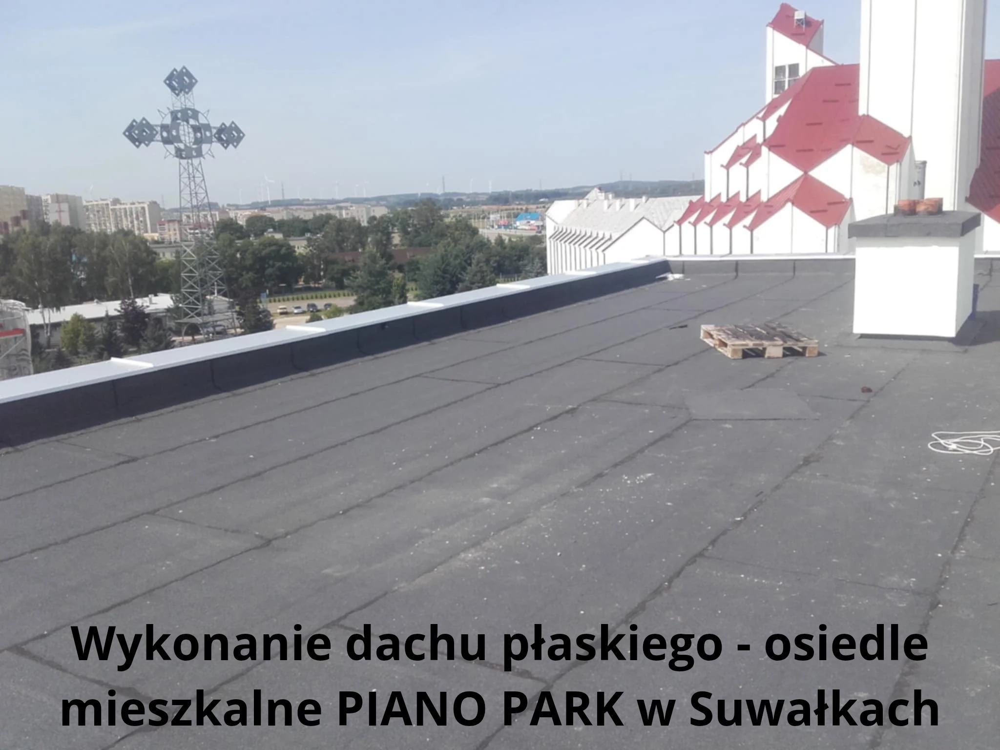 Wykończony płaski dach z widokiem na miasto i kościół w Suwałkach. Paleta leży na dachu, a w tle widoczne turbiny wiatrowe.